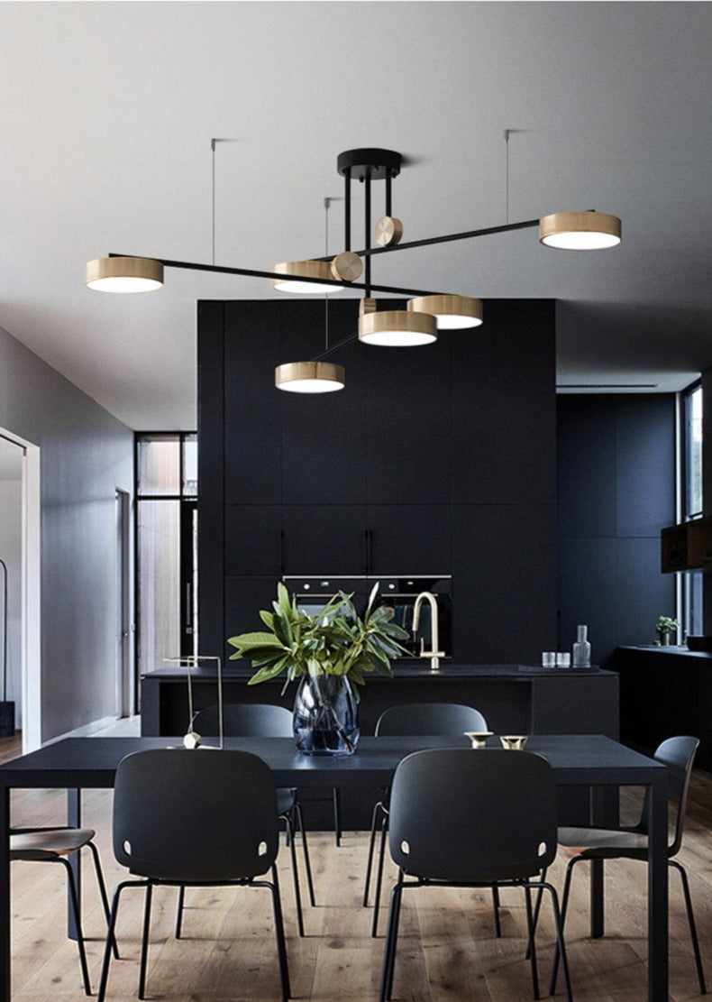 Modern pendant rotating chandelier with a black canopy and gold-colored metal lights, hanging over a dark dining area with a black table and black chairs.